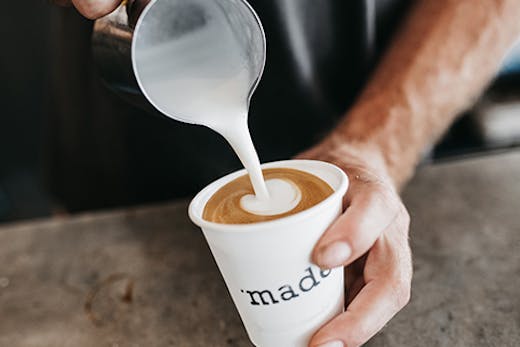 A close-up of two hands holding a white coffee cup that says 'made' on it while pouring frothing milk in the cup.