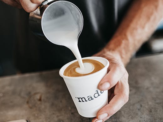 A close-up of two hands holding a white coffee cup that says 'made' on it while pouring frothing milk in the cup.