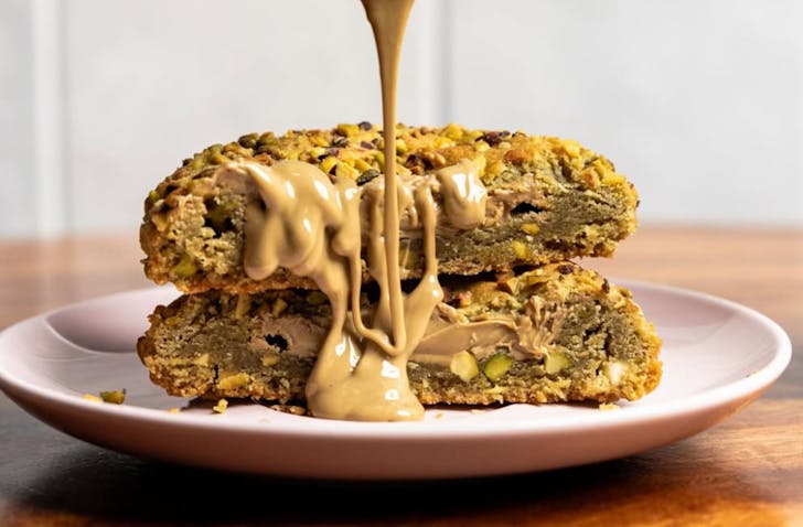 A close up on Kurtosh's Princess Fiona cookie cut in half. The image shows the creamy, green interior of the cookie, and the crumbly and nutty outside. A light drizzle of pistachio cream is being poured onto the cookie by an outside-of-frame hand.