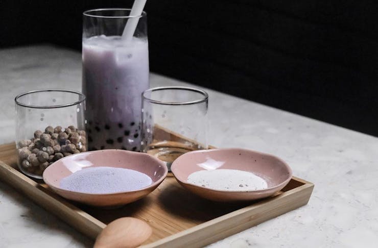 A purple bubble tea in a glass beaker, on a tray with two bowls of pink and purple tea powder