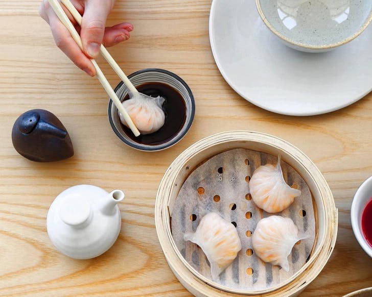 Above view of steamed dumplings in a bamboo steamer. A hand is holding chopsticks and has grabbed one of the four dumplings, now dipping it in sauce.