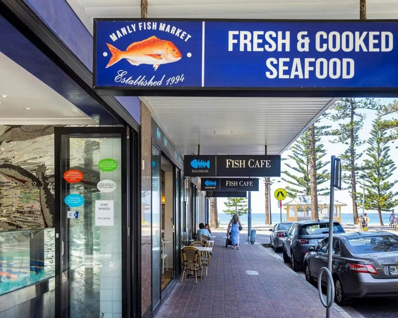 Outside view of the Manly Fish Market store sign, with the photograph taken from the point of view of a person walking down the pavement sidewalk.