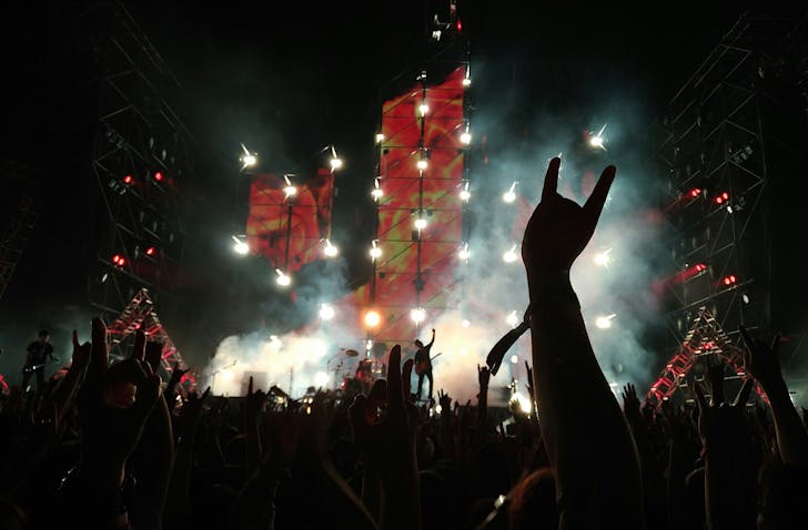 Silhouetted rock ravers hold their hands up during a concert, closest to the camera, someone gives the heavy metal horn symbol with their hand. 