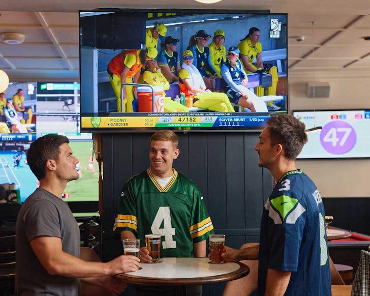 Three men sit around a pub table chatting while a screen on the wall above and behind them plays the live sports game. The men are dressed in jerseys and holding drinks such as beer.