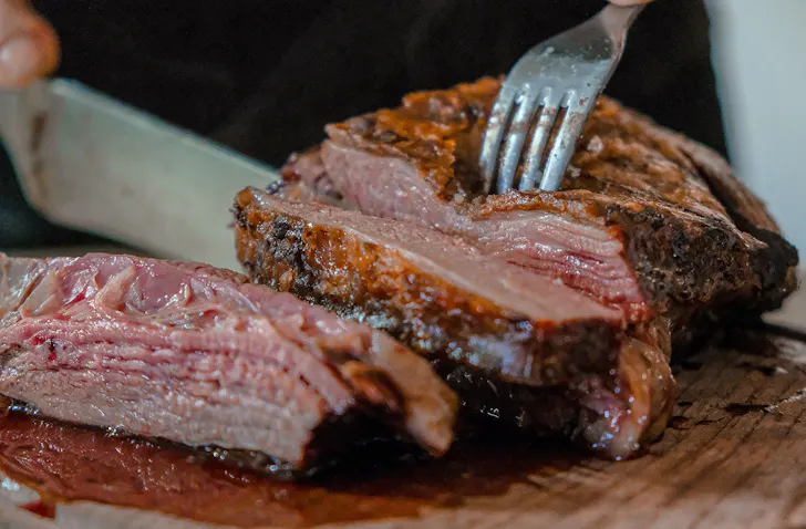 A hand cutting a piece of smoked BBQ meat with fork and knife