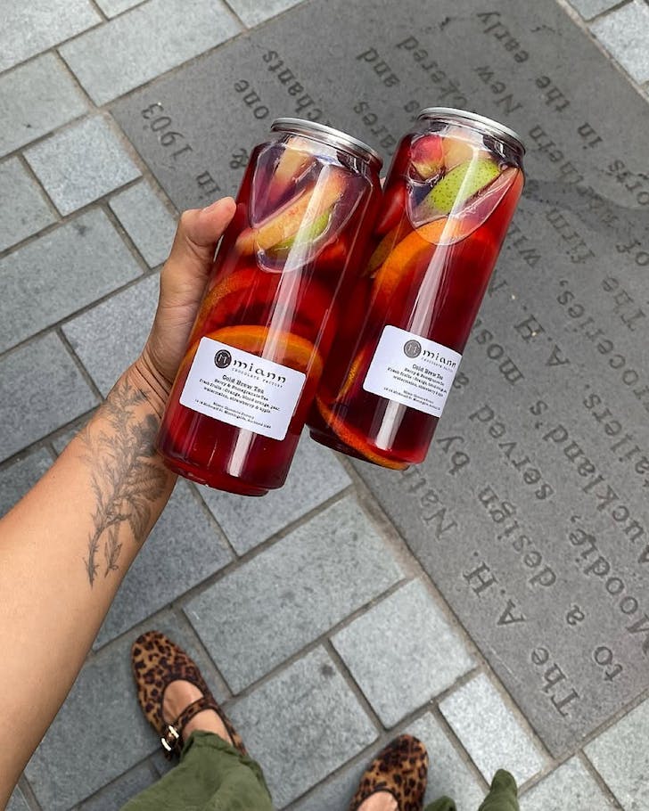 Someone with a gorgeous botanical forearm tattoo holds two cans of cold brew tea stuffed with summer fruit.