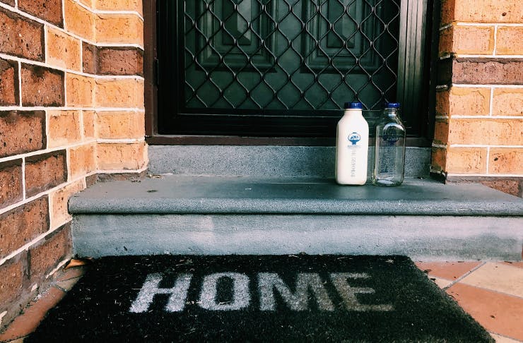 A glass bottle filled of milk sitting on a doorstep. A doormat sits in front with the words 