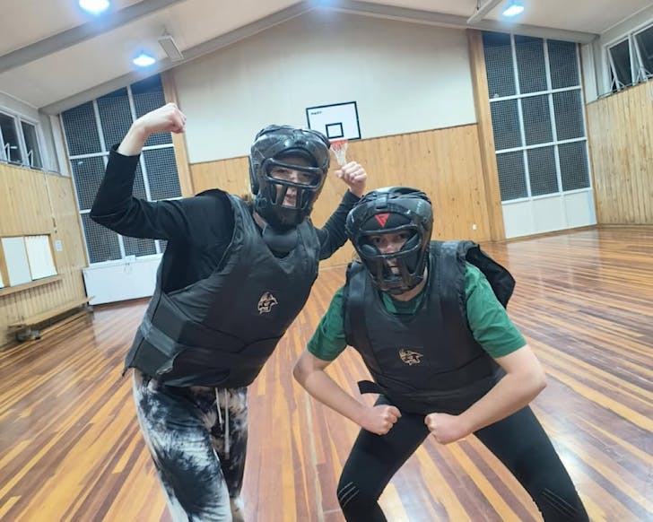 Two women standing in a gymnasium in padded protective gear flex their muscles.