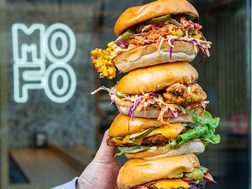 Three burgers stacked on top of each other in front of a neon sign that reads 'Mofo