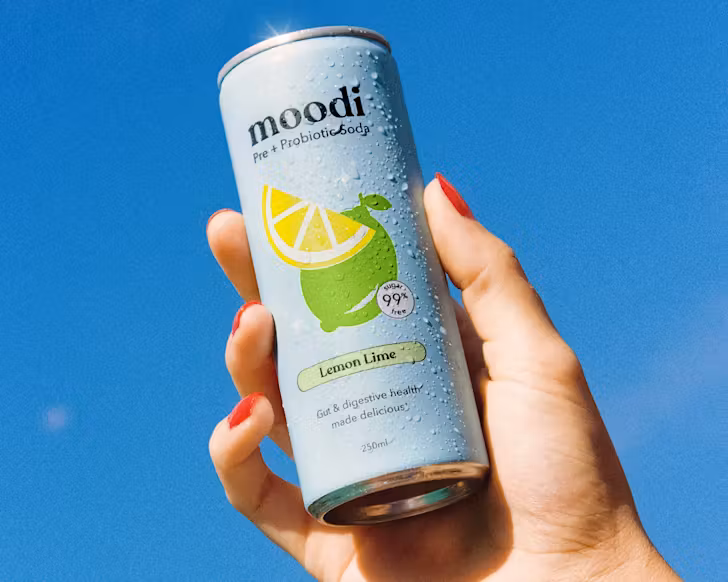 Someone with red nail polish holds a light blue can of pre + probiotic soda up to the deep blue sky. 