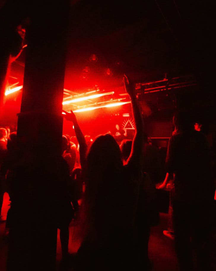 A dark red-lit rave room with a glowing Morning People logo and red lights in front and above the crowd, respectively.