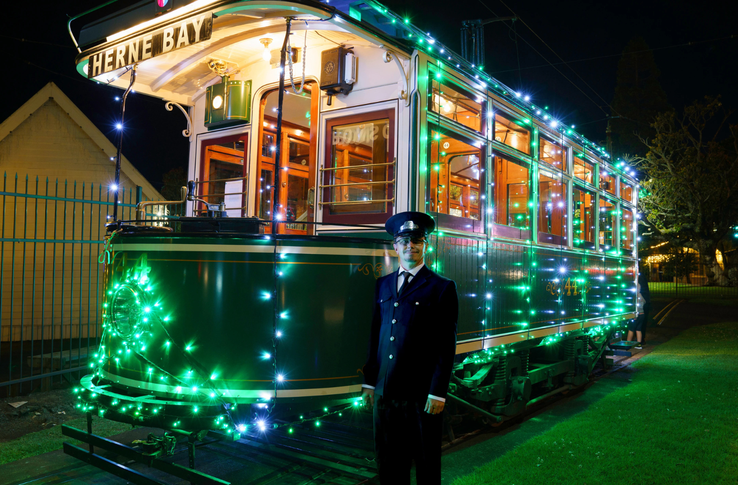 A MOTAT conductor stands before an old green tram carriage adorned in green festive lights.