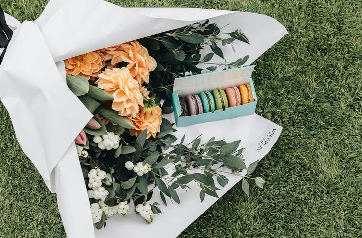 A bouquet of flowers wrapped in white paper sitting on green grass with a box of colourful macarons 