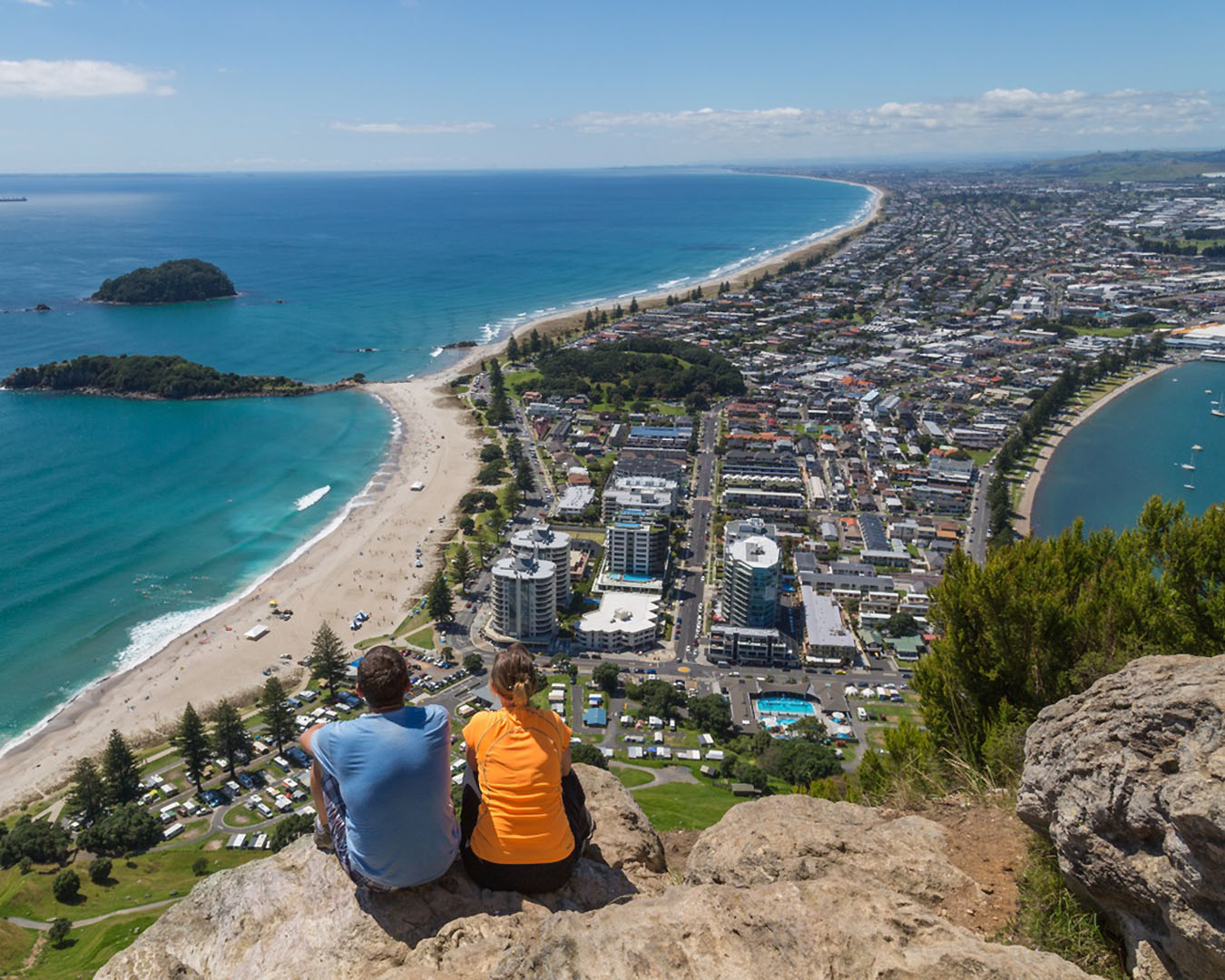 Two people sit at the summit of Mauao and look down on Mount Maunganui.