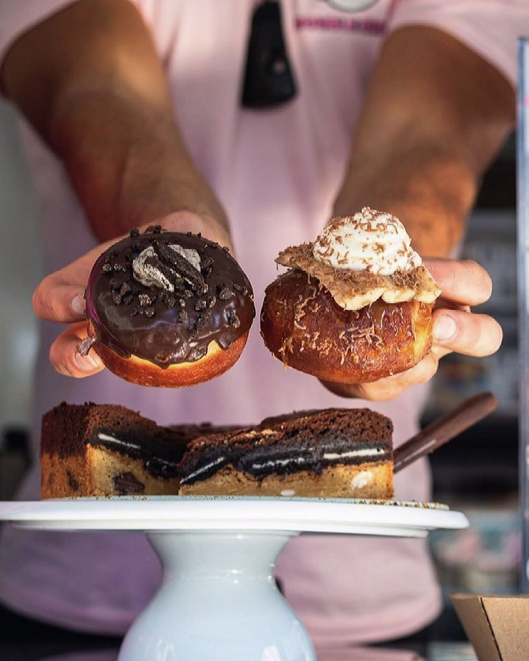Someone holds up amazing-looking doughnuts with a cake in the foreground.