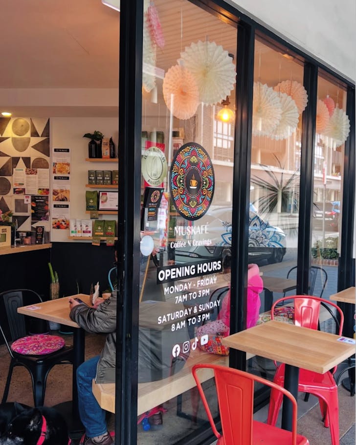 An mid-shot image of the outside of Muskafe where there are colourful decor chairs and a large window where we can see more colour inside, as well as people sitting and eating.