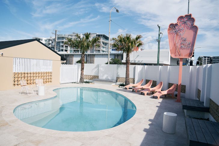 the exterior of a retro motel with kidney shaped pool and a neon pink sign and a palm tree