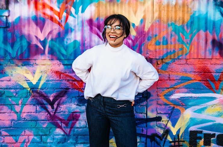 A girl with glasses smiling in front of a bright graffitied wall. 