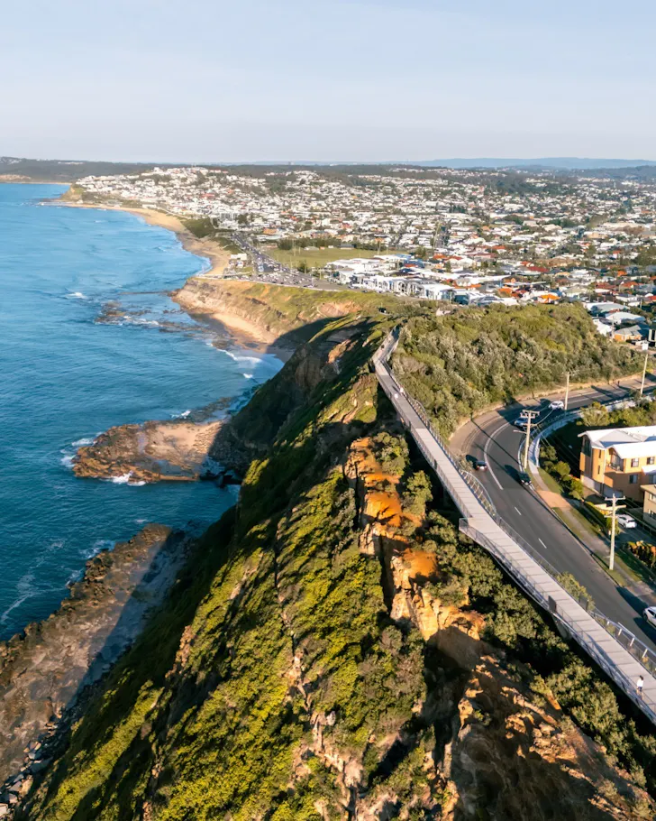 Aerial view of a rugged coastal cliff lined with a pedestrian boardwalk overlooking the ocean, with waves below and the suburban coastline stretching into the distance.