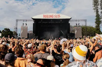 Crowd gathered in front of the main Rolling Sets festival stage, with hands raised as a band performs beneath a bright blue sky framed by scattered clouds and tall pine trees.