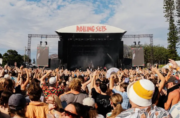 Crowd gathered in front of the main Rolling Sets festival stage, with hands raised as a band performs beneath a bright blue sky framed by scattered clouds and tall pine trees.