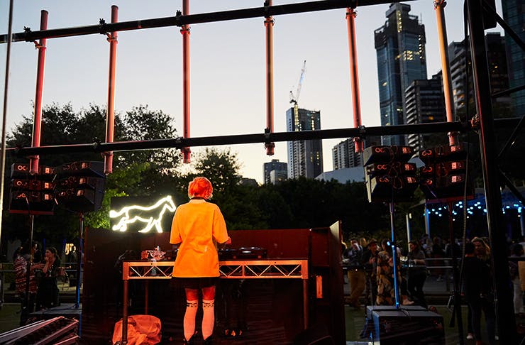 A DJ playing to a large crowd outside the NGV at night.