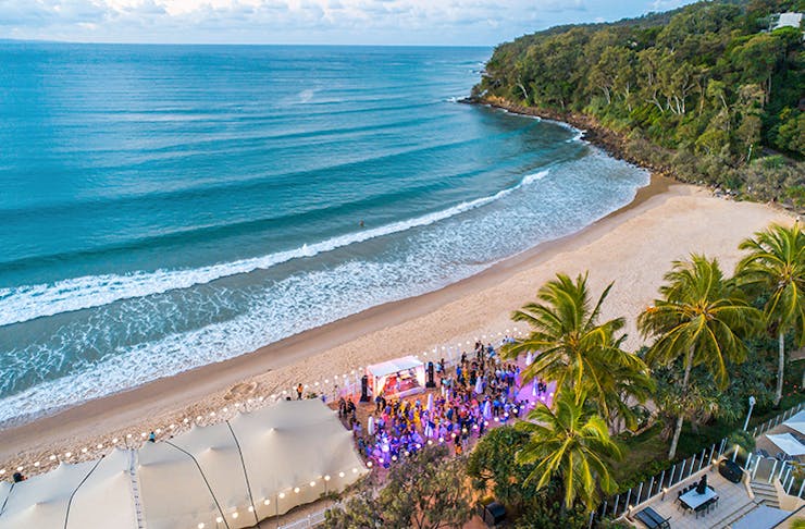 An aerial shot of a crowd on a beach under a white canopy of tents and fairy lights.