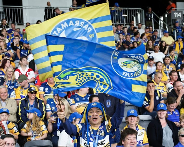 A medium-shot photo of the crowd at a Parramatta Eels NRL game, with many people wearing the jerseys of this team. A man at the forefront of the image is standing and waving two Parramatta Eels flags.