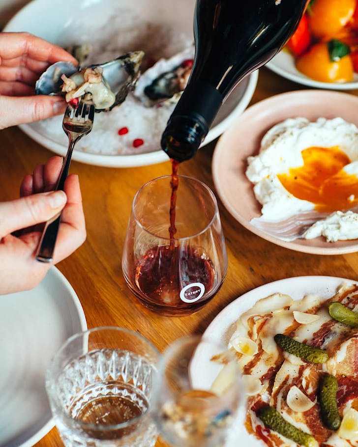 A server pours wine, while someone scoops out an oyster while enjoying plates of delicious food from Odettes Eatery.