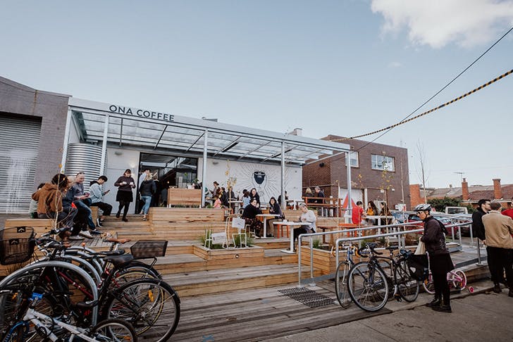 A warehouse cafe drenched in sunlight. Cyclists sit out the front with parked bikes in bicycle lock-ups.