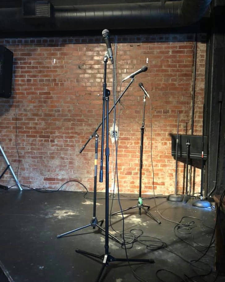 A bunch of mics on stands on an empty stage with a brick back wall.
