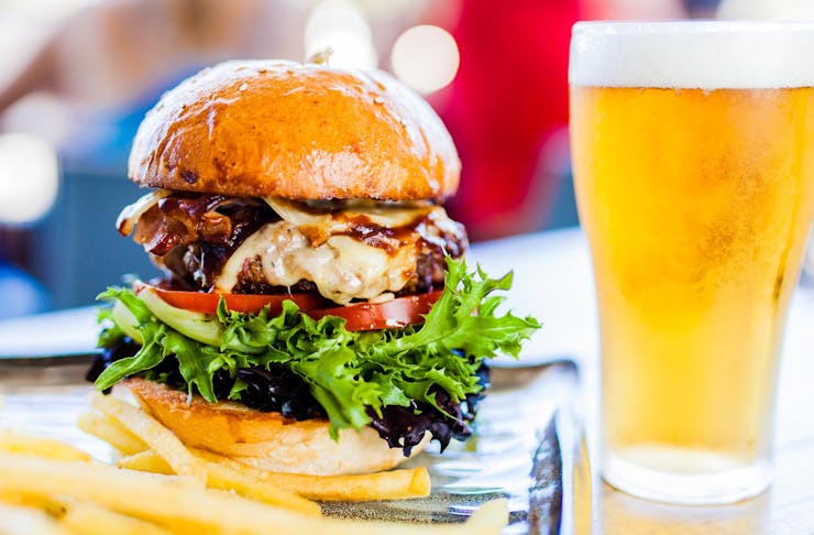 A deliciously-looking burger piled high with a meat patty, cheese, tomato and lettuce, sits alongside of a beer in a schooner glass.