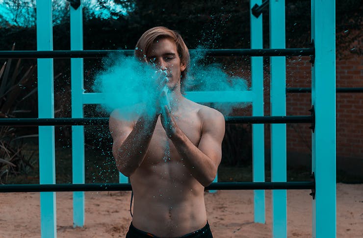 A man claps his hands together with a puff of blue chalk, he stands in front of blue work out bars.
