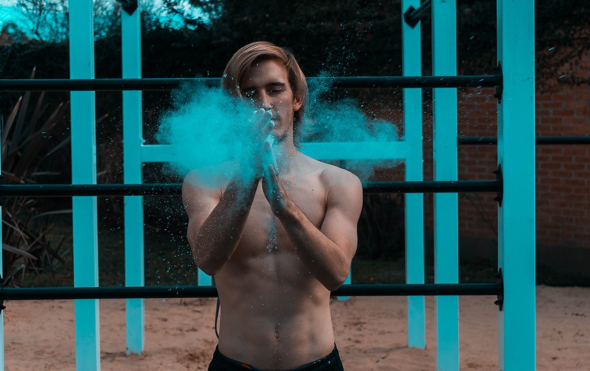 A man claps his hands together with a puff of blue chalk, he stands in front of blue work out bars.