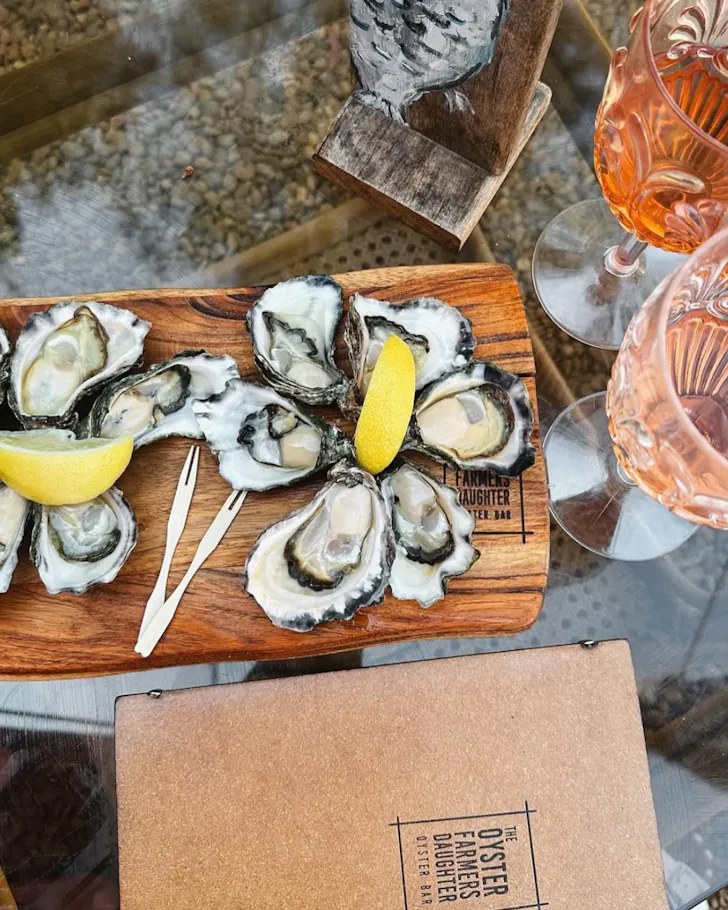 A board full of fresh oysters with lemon wedges and two glasses of rose at The Oyster Farmers Daughter.
