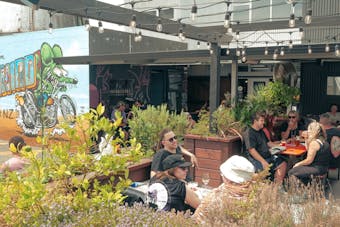 Wide shot showing the outdoor dining area of Panhead Brewery at Brewtown. There is artwork on the outer wall of the brewery and many people sitting on outdoor tables drinking and chatting during the daytime.