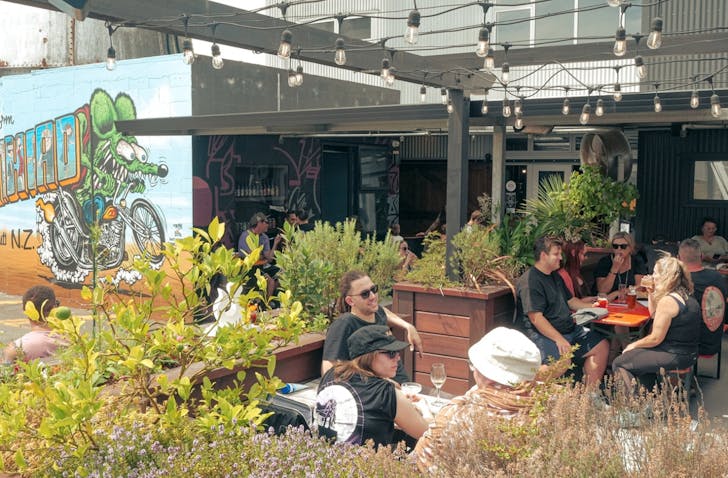 Wide shot showing the outdoor dining area of Panhead Brewery at Brewtown. There is artwork on the outer wall of the brewery and many people sitting on outdoor tables drinking and chatting during the daytime.