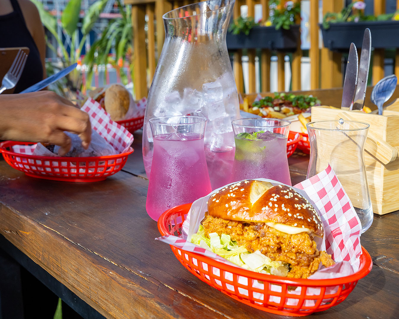 A burger sits in the foreground with a jug of cocktails standing behind it while someone tucks in.
