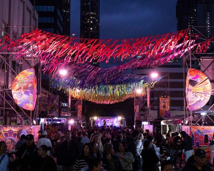 A wide shot of an outdoor Parramatta street that has been decorated with colourful streamers and hanging decor and lights. There are crowds of people walking around and visiting stores shown at the side of the image. A music stage is shown at the end of the street, with colourful lights and screens on display.