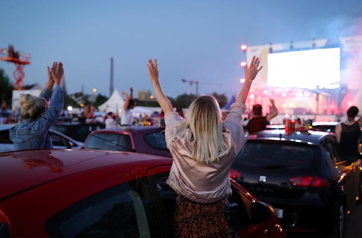 A woman with her hands raised, facing away from the camera towards a stage and a carpark full of cars and other people.