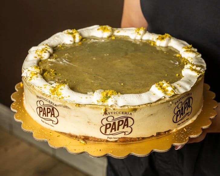 An image of a round pistachio tiramisu cake, that has green cream on top and is wrapped with a cake paper that has 'Pasticceria Papa' logo on it.
