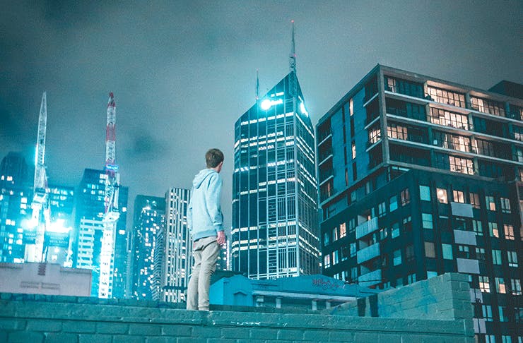 A man stands atop a wall looking at the Melbourne city skyline at night.