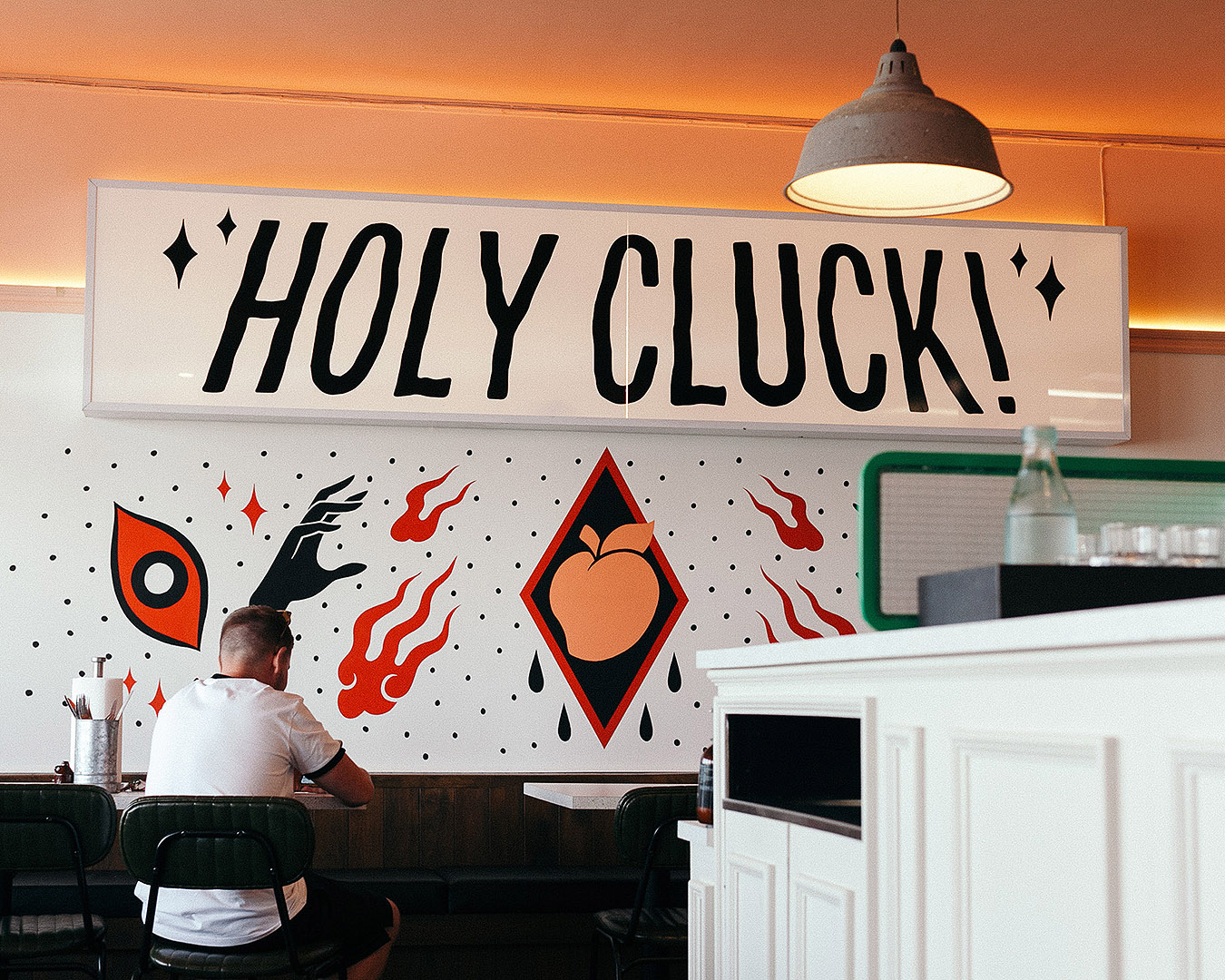 We’re looking at the wall of a restaurant at ‘HOLY CLUCK’ in large lettering above an artwork with single eyes, a peach in a black triangle, fire and black arms. 