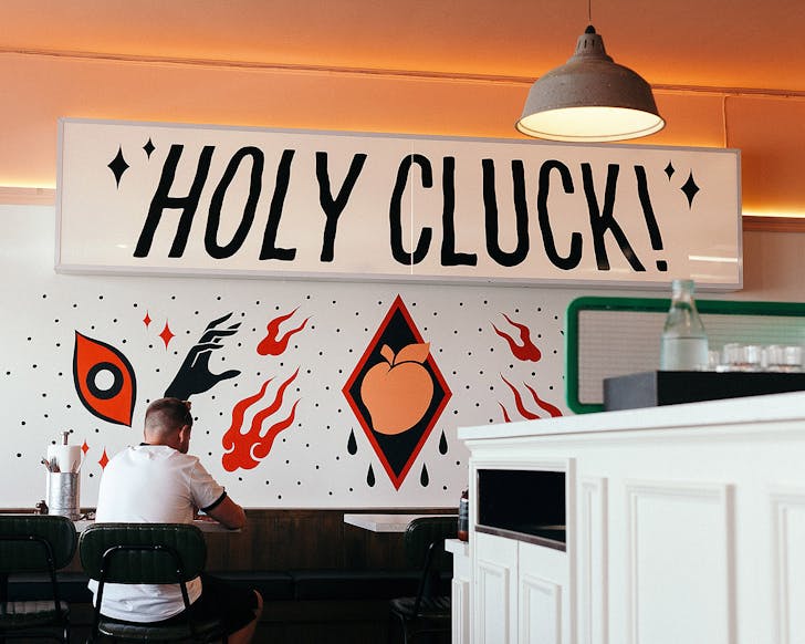 Weâre looking at the wall of a restaurant at âHOLY CLUCKâ in large lettering above an artwork with single eyes, a peach in a black triangle, fire and black arms. 