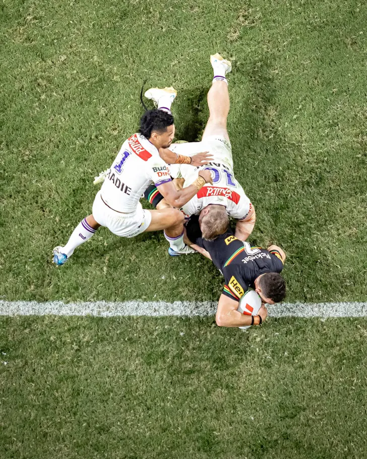 Rugby league player scores a try on the line as two defenders attempt a last-ditch tackle.