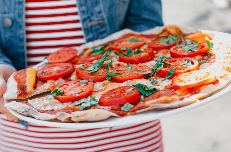 A woman holds a delicious thin crust pizza with huge tomatoes on top.