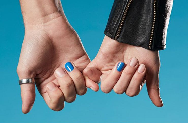 Two hands in front of a blue background. Both pinky fingers are interlocked, while the nails on the ring fingers are painted blue.