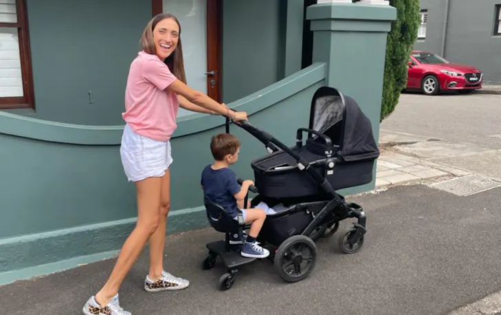 Lady smiling as she takes her kids for a walk in a pram along a street. 