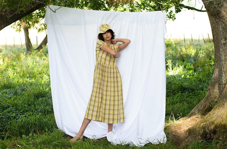 Woman in a yellow gingham dress and bucket hat stands in front of a sheet in a field. 