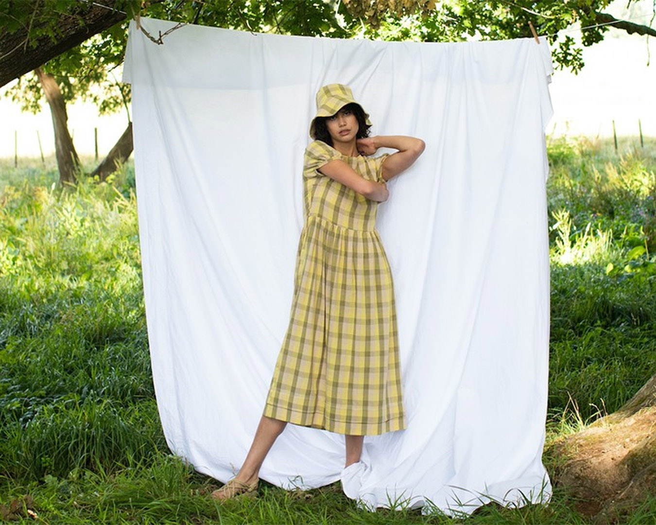 Woman in a yellow gingham dress and bucket hat stands in front of a sheet in a field. 